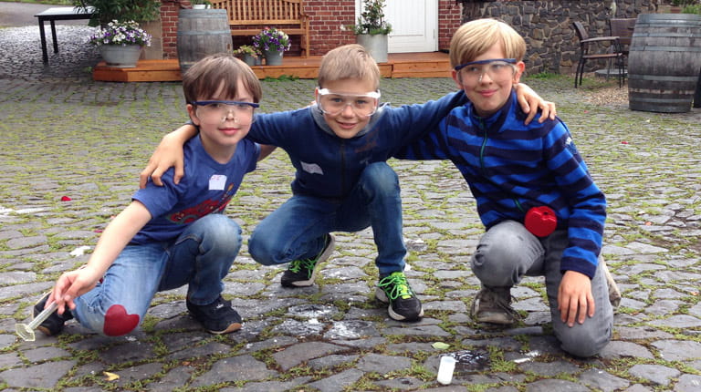 Drei Kinder knien auf einem Hof und tragen Schutzbrillen.