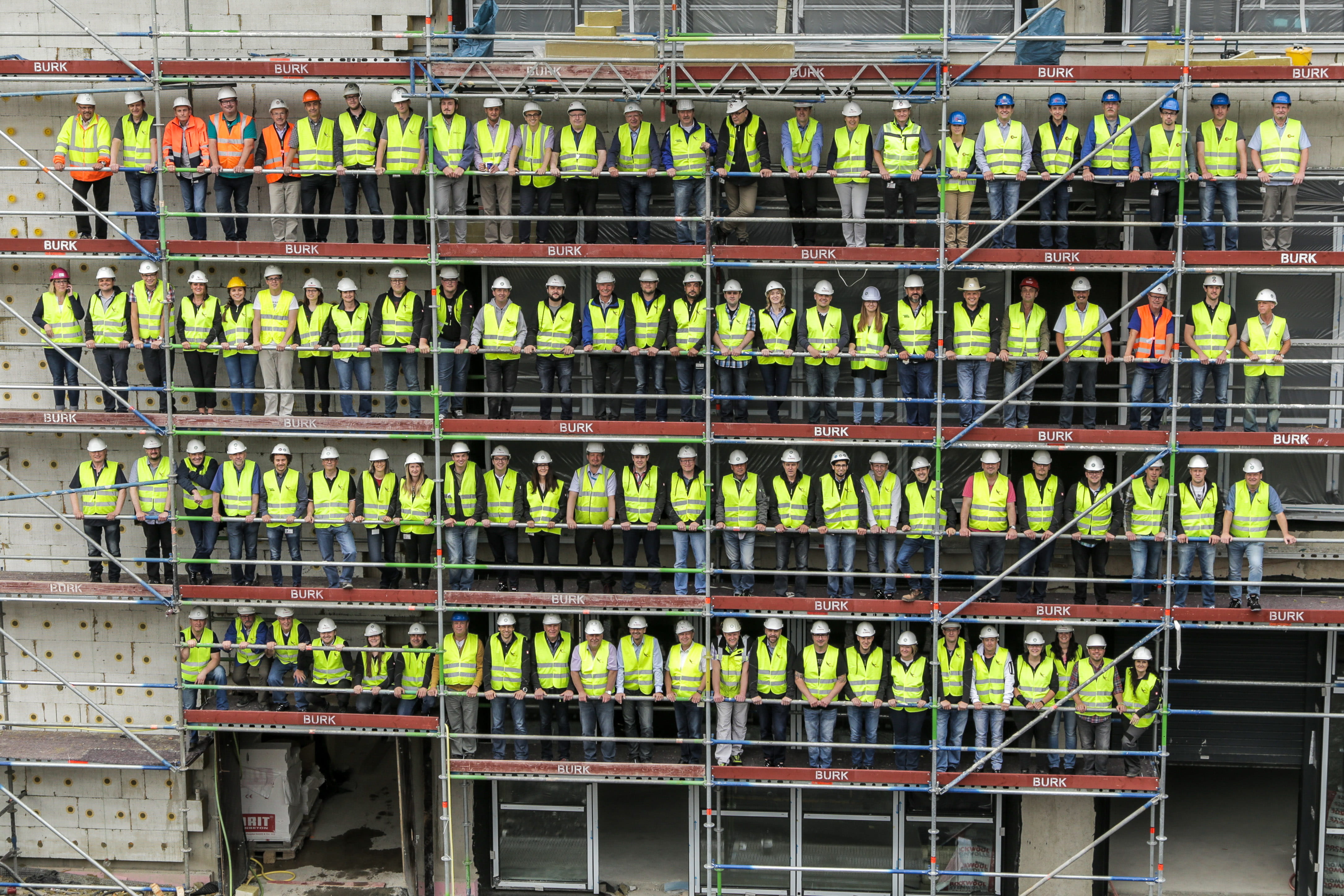 99 Menschen in gelben Westen, mit Bauhelmen auf Baugerüst