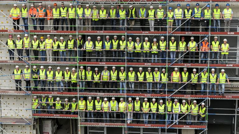 99 Menschen in gelben Westen, mit Bauhelmen auf Baugerüst