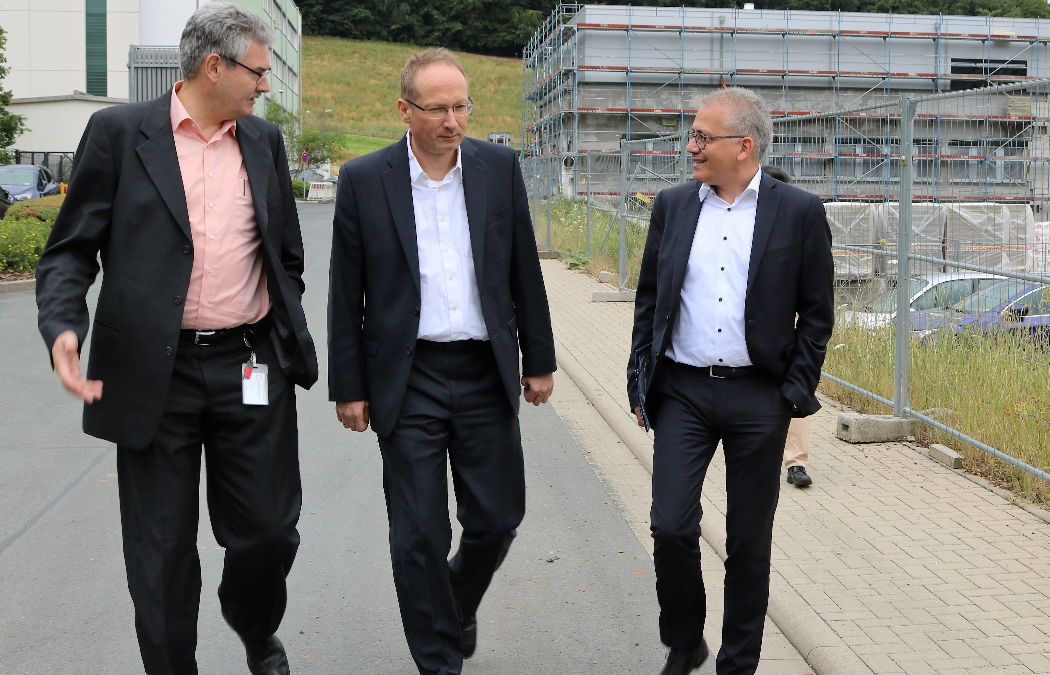 Drei Männer in Anzügen unterhalten sich und gehen an Baustelle vorbei.