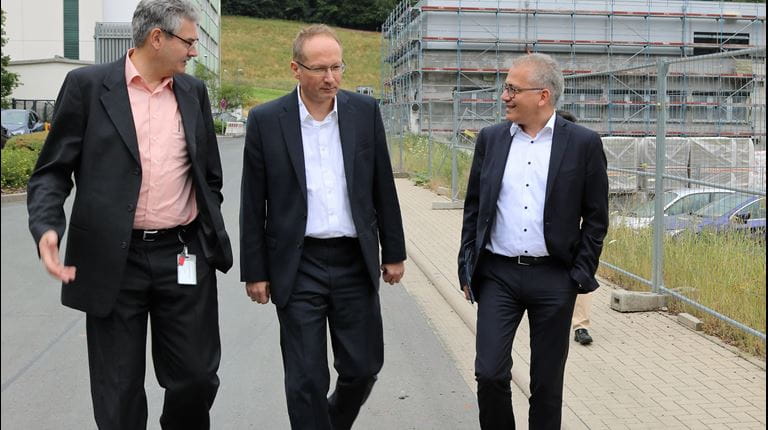 Drei Männer in Anzügen unterhalten sich und gehen an Baustelle vorbei.