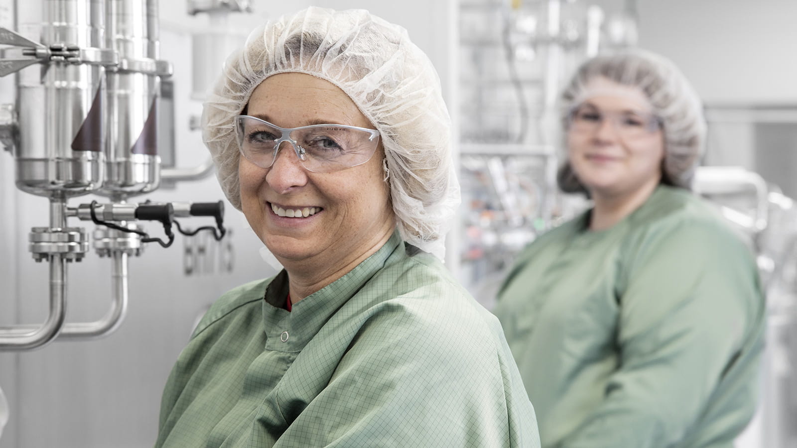 Zwei Frauen in Produktionsumfeld mit Overalls, Haarnetz und Schutzbrillen lächeln.